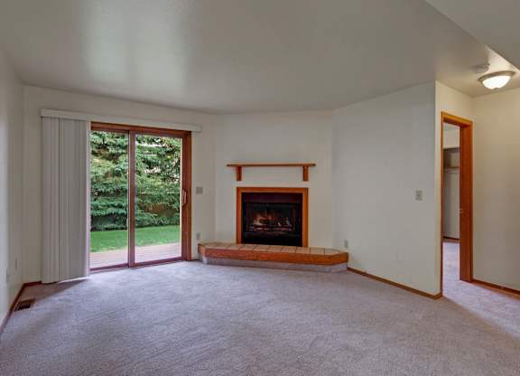 A spacious, empty living room here at Wildwood Estates with soft carpet, a cozy fireplace with a tiled hearth, and a sliding glass door that opens to a patio with peaceful green views.