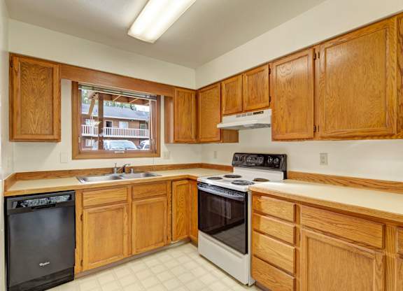 A bright kitchen here at Wildwood Estates with warm wood cabinets, light countertops, a stainless steel sink, and a dishwasher, featuring a stove and a large window that brings in natural light.