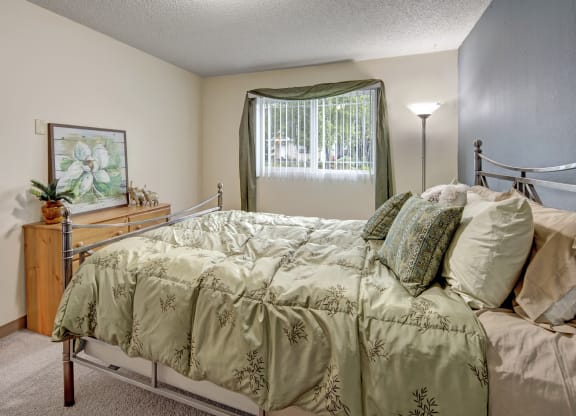 Cozy bedroom here at Brighton Wood Apartments with a green bedspread and matching pillows, a wooden dresser with decorative items, and a window with green curtains, creating a serene vibe.