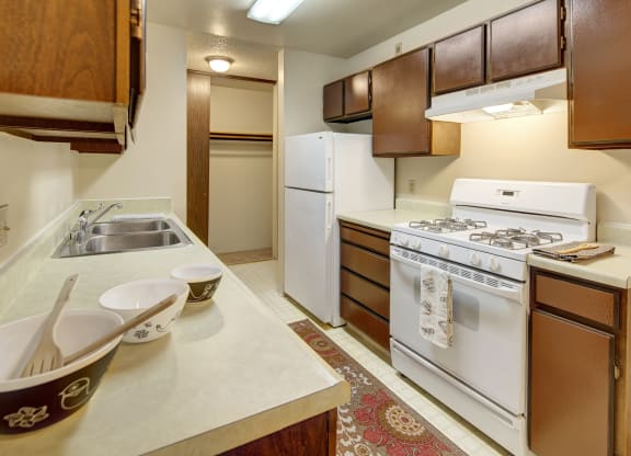 Narrow kitchen here at Taiga Apartments with brown cabinets, white appliances including a refrigerator and gas stove, three bowls on the counter, and a patterned rug on the floor.