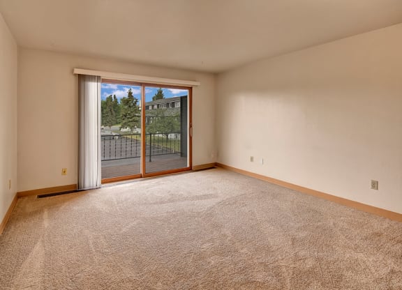 An empty living room with beige carpet and white walls. A large sliding glass door opens to a balcony with a view of trees and buildings, conveying a serene atmosphere. The Alaskan Apartments.