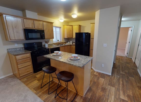 Bright kitchen here at Four Seasons Apartments with wooden cabinets, granite countertops, and a black appliance set. Two wooden stools at an island create a cozy, welcoming ambiance.
