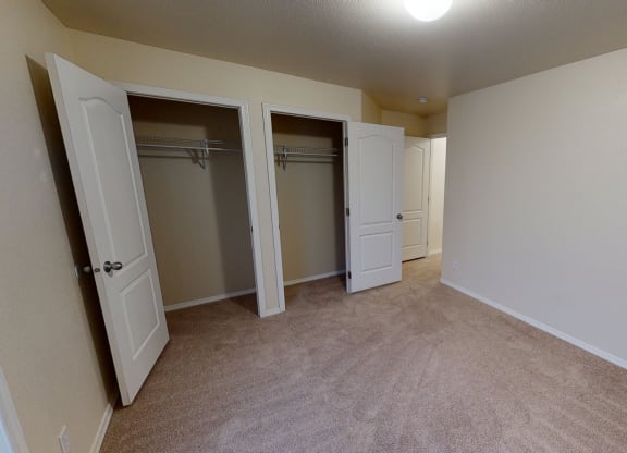 Bedroom here at Four Seasons Apartments with beige carpet and cream walls, featuring two open closets with white double doors, creating a spacious and neutral atmosphere.
