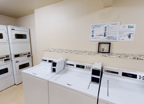 A clean laundry room here at Hillside Chalet Apartments with stacked dryers on the left and top-loader washers on the right. Instructions are mounted on a beige wall, creating an orderly atmosphere.