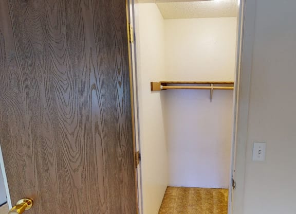 Open closet here at Rancho Tudor Apartments with wooden door ajar, revealing a white interior, a wooden hanging rod, and beige carpet. The space is minimal and tidy.