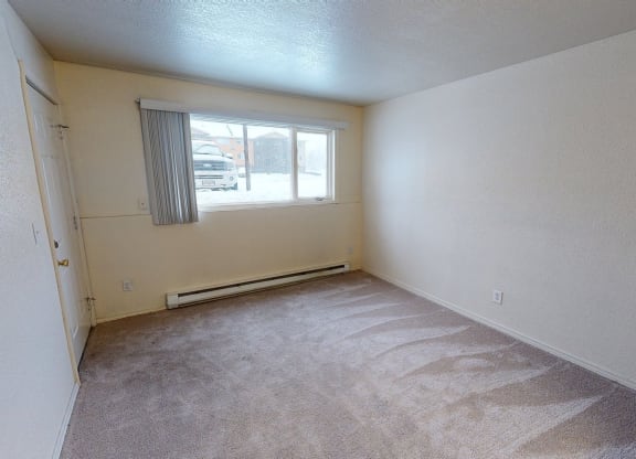 Empty living room here at The Club at Eagle Pointe Apartments with beige walls and carpet, featuring a large window with vertical blinds. Sunlight highlights the carpet's texture, creating a calm atmosphere.