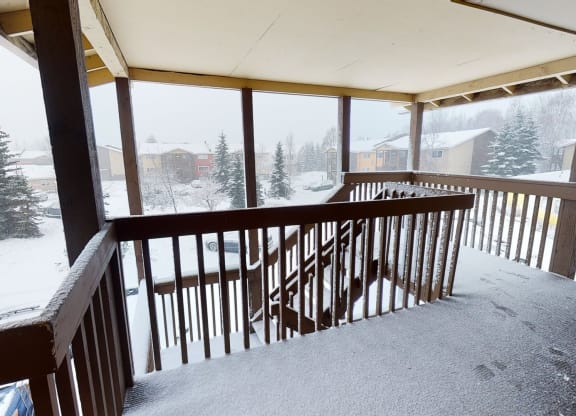 Snow-covered porch here at The Club at Eagle Pointe Apartments with wooden railings overlooks a snowy neighborhood. Fir trees and houses are visible under a gray, overcast sky. Quiet, wintry scene.