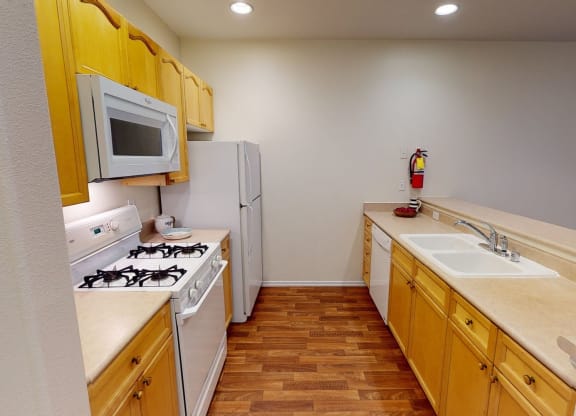 Compact kitchen here at The Highlands Luxury Apartments with wooden cabinets, white appliances, and beige countertops. Gas stove, microwave, and sink are visible. Cozy and functional atmosphere.