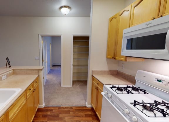 Kitchen interior here at The Highlands Luxury Apartments with light wood cabinets, a gas stove, white microwave, and double sink. Hallway with open pantry on the right leads to another room.