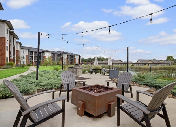 Outdoor patio here at 49 West Apartments with Adirondack chairs around a square fire pit. String lights hang above a pathway, leading to modern apartments and lush greenery. Relaxing ambiance.