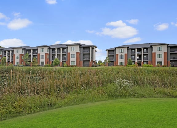 Modern three-story 49 West Apartment buildings with a mix of beige and brick exteriors sit behind a lush wetland, under a clear blue sky.