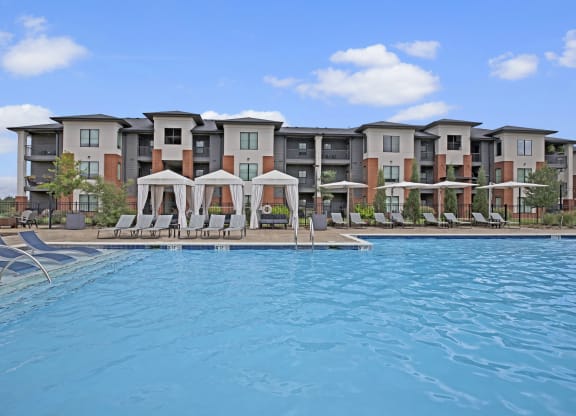 Luxurious poolside view of 49 West's modern apartment complex, featuring cabanas and lounge chairs under a blue sky with fluffy clouds. Relaxing and inviting atmosphere.