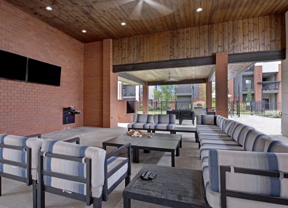 Outdoor lounge area here at 49 West Apartments with striped blue and white cushioned chairs and tables under a wooden ceiling. Two wall-mounted TVs; relaxed and inviting atmosphere.