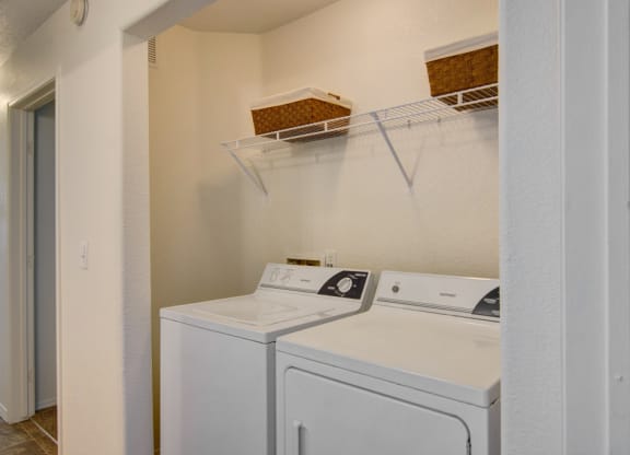 Laundry area here at Arezzo Apartments with a washer and dryer, topped with a metal shelf holding two brown baskets. The space is clean and organized, with a simple white interior.