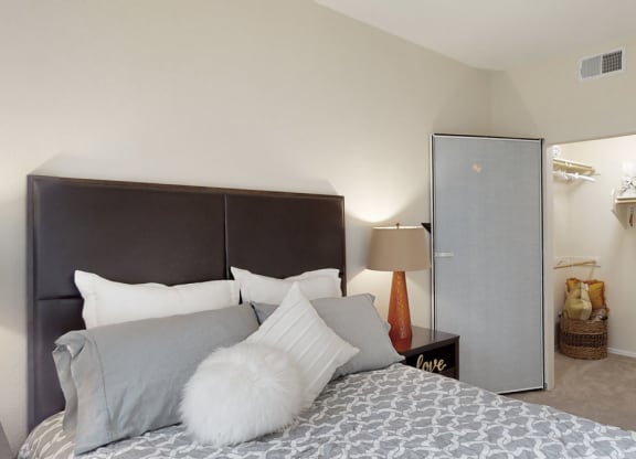 Bedroom interior here at Acclaim Apartments with a neatly made bed, decorative pillows, and a dark headboard. A modern lamp on a side table, and an open closet with baskets conveys a cozy, organized atmosphere.