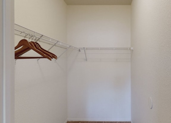 Empty, light beige walk-in closet here at Arezzo Apartments with a wire shelf on the left wall holding a few wooden hangers. Carpeted floor gives a minimalist feel.