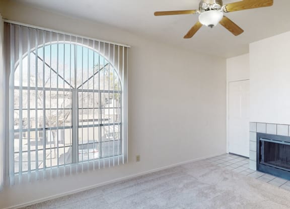 Bright living room here at Desert Horizon Apartments with a large arched window with vertical blinds, beige carpet, white walls, a ceiling fan, and a black-tiled fireplace.
