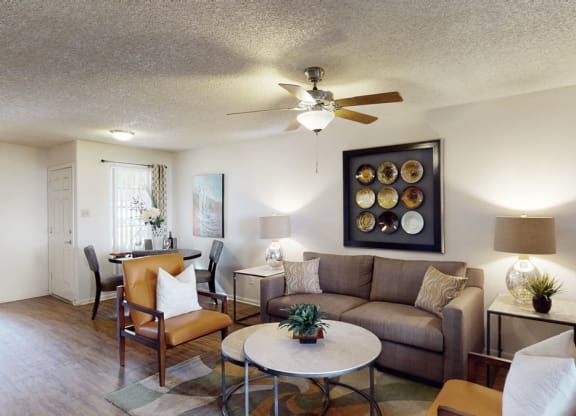 Living room here at Desert Horizon Apartments with a cozy beige sofa, two brown chairs with white pillows, and a round coffee table with a plant. Decorative wall art features nine plates. There are two side tables with lamps, and a dining area with a table and chairs is visible in the background. Ceiling fan and soft lighting create a warm, welcoming atmosphere.