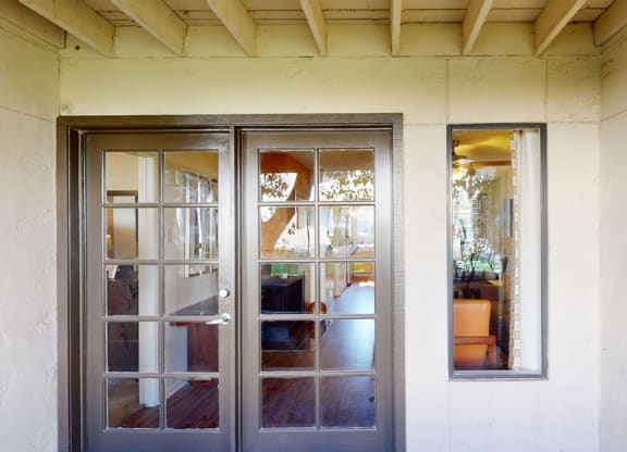 Glass double doors here at Desert Horizon Apartments and a window framed in brown open to a bright interior, reflecting outdoor trees. Simple, inviting entryway with neutral tones.