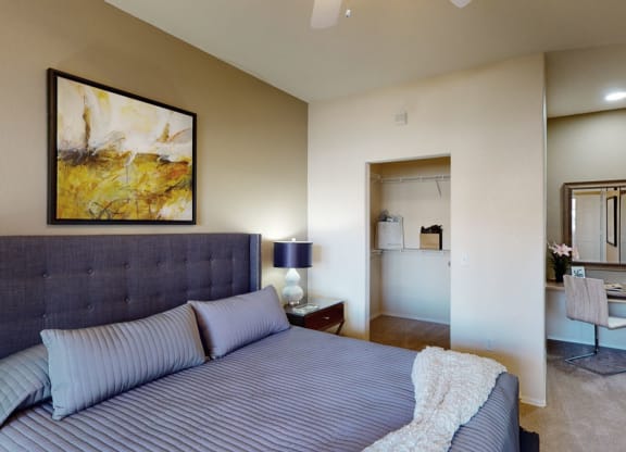 Cozy bedroom here at Dobson Towne Centre Apartments with a gray upholstered bed, striped bedding, and abstract art. A lamp and nightstand are beside the bed. A closet and vanity area are visible.