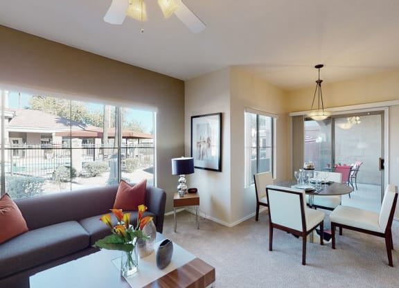 Modern living room here at Dobson Towne Centre Apartments with large windows overlooking a pool area. Gray sofa with orange pillows, glass-topped table with flowers, and dining area with white chairs. Bright and inviting atmosphere.