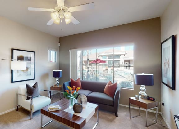 Contemporary living room here at Dobson Towne Centre Apartments with gray sofa, white armchair, and wood-accented furniture. Large windows overlook a patio. Neutral tones, cozy ambiance.