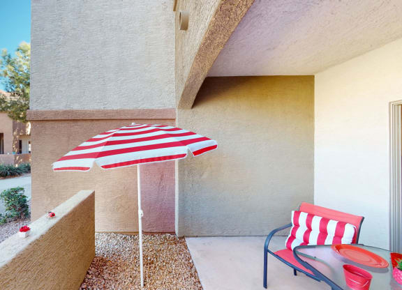 A sunlit patio here at Dobson Towne Centre Apartments features a red and white striped umbrella and matching chair cushion. Nearby, a glass table holds red items. A house and tree are visible in the background, conveying a cozy, cheerful vibe.