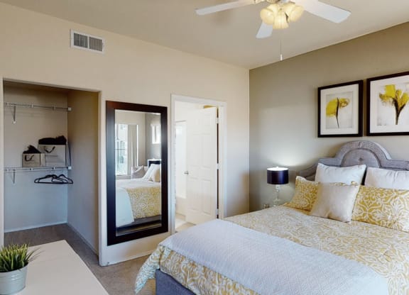 Bedroom here at Dobson Towne Centre Apartments with a stylish gray bed, patterned yellow bedding, and two framed yellow artwork pieces above. A mirrored closet reflects a calm, cozy ambiance.