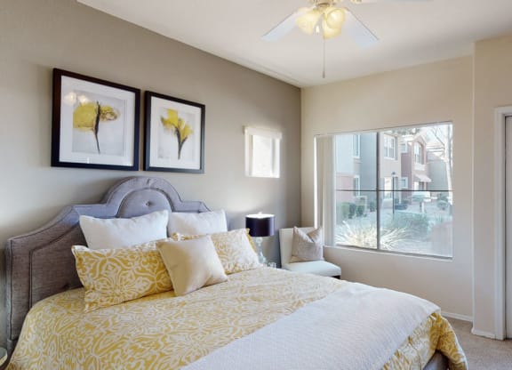 Bright bedroom here at Dobson Towne Centre Apartments with a gray upholstered bed, yellow-patterned bedding, and floral art. Sunlit window and door open to an outside view, creating an airy feel.