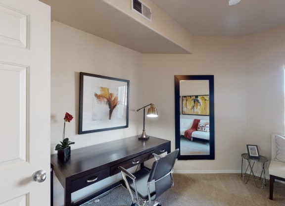 A modern office space here at Dobson Towne Centre Apartments with beige walls features a dark wood desk, a sleek lamp, and a reflective mirror. There's a gray chair, abstract art, and a small plant on the desk, creating a calm and organized atmosphere.