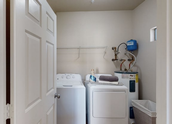 A small laundry room here at Dobson Towne Centre Apartments with a washing machine and dryer. A shelf above holds a detergent bottle and folded towels. A water heater is to the right.
