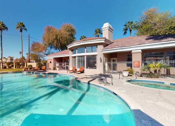 Outdoor pool area here at Dobson Towne Centre Apartments with clear blue water, surrounded by palm trees and sun loungers. Adjacent is a beige building with large windows under a sunny blue sky.