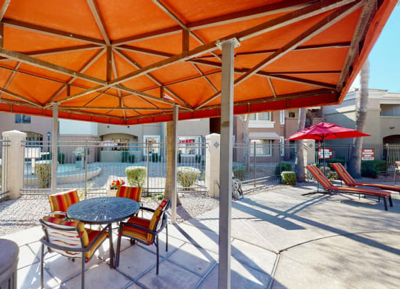 Outdoor patio here at Dobson Towne Centre Apartments under an orange canopy with a round table and striped chairs. Nearby, pool loungers with red umbrellas. Sunny and inviting ambiance.