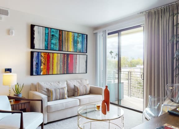 Modern living room here at Escape Apartments with abstract art, cream sofa, and striped cushions. A glass coffee table holds a red vase and glass. Large window with beige curtains reveals a balcony and palm trees outside. Bright and inviting.