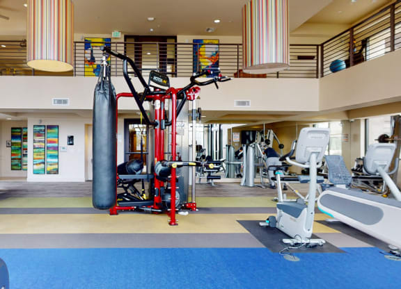 Spacious gym interior with modern equipment here at Escape Apartments, including a red multi-station machine and a punching bag. Brightly colored abstract art decorates the walls.