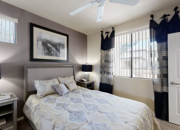 A cozy bedroom here at Natura Villas Apartments with a neutral color palette. Features a bed with patterned bedding, two bedside tables with lamps, abstract wall art, and window with curtains.