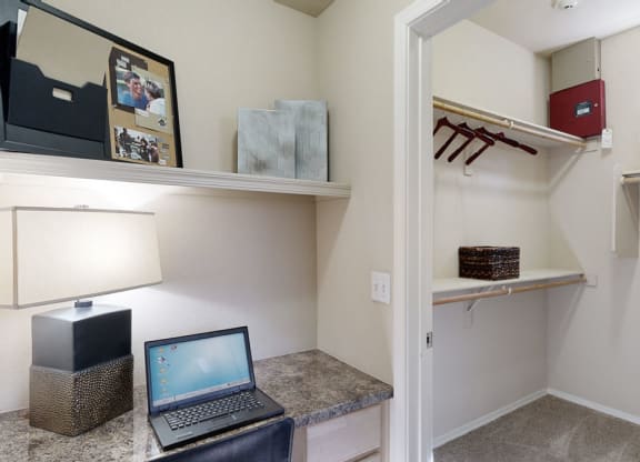 Cozy nook with a laptop on a granite desk here at Natura Villas Apartments, lit by a modern lamp. Nearby, an open closet shows hangers and a wicker basket on shelves.