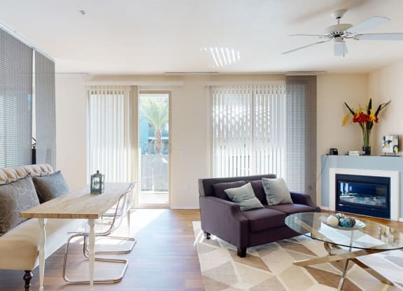 Modern living room here at Rio Paradiso Apartments with natural light, featuring a gray sofa, glass coffee table, dining bench, and decorative fireplace. Minimalist and cozy atmosphere.