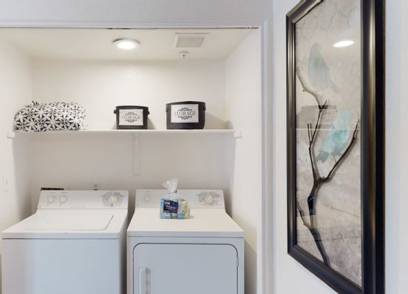 Laundry nook with a washer and dryer here at The Hawthorne Apartments, a shelf above holds patterned bins and a black-and-white laundry bag. A framed bird artwork is on the right wall.