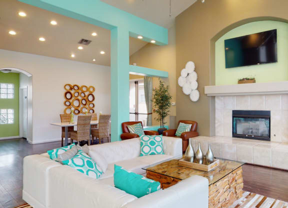 A modern living room here at The Hawthorne Apartments with teal accents features a white, L-shaped sofa with teal cushions, a fireplace, wall art, and a TV. A dining area is visible in the background.