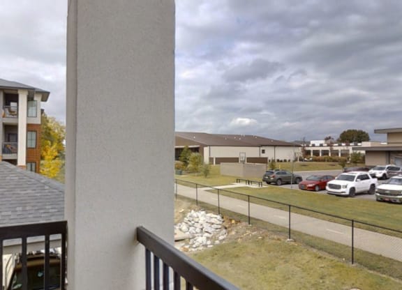 View from a balcony here at 49 West Apartments overlooking a parking lot with several cars, a fenced pathway, and nearby modern buildings under a cloudy sky, conveying a calm, suburban atmosphere.
