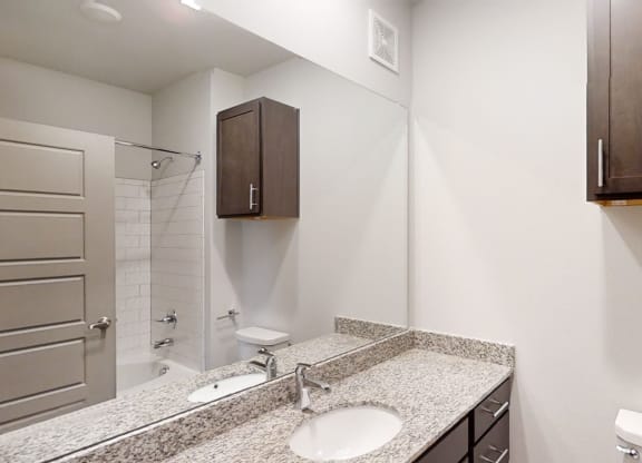 Modern bathroom here at 49 West Apartments with a granite countertop, single sink, dark wooden cabinets, and a white tiled bathtub. The space is clean and minimalistic.