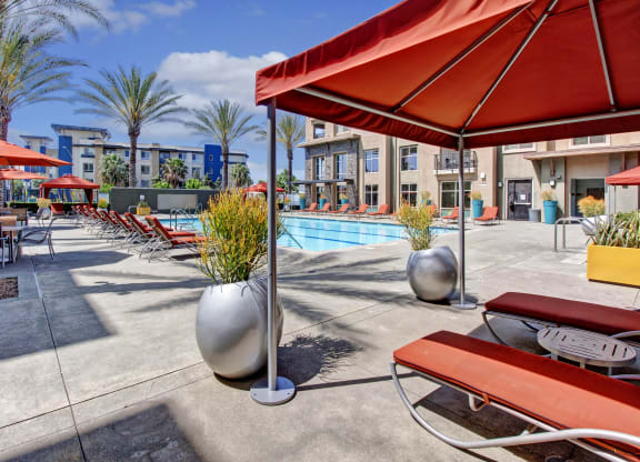 Outdoor pool area here at Artisan at Main Street Metro Apartments with red lounge chairs and umbrellas, surrounded by palm trees and modern buildings under a clear blue sky. Relaxing ambiance.