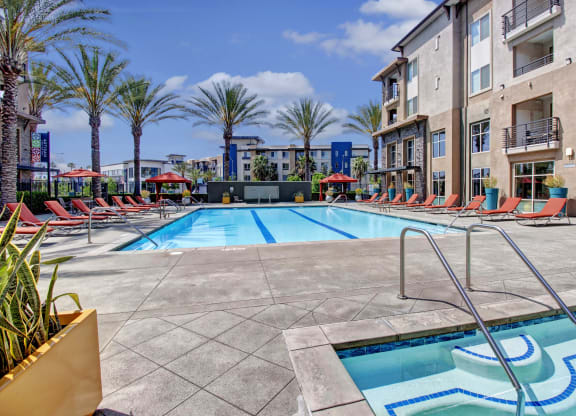 Artisan at Main Street Metros modern apartment complex pool area with red lounge chairs, palm trees, and umbrellas under a blue sky. A hot tub is in the foreground. Bright and inviting.