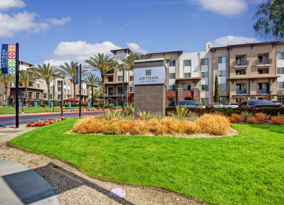 Modern apartment complex with lush green lawn in foreground. Sign reads “Artisan at Main Street Metro.” Palm trees and colorful banners line the street, under a blue sky.