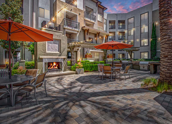 Cozy outdoor patio here at Artisan at Main Street Metro Apartments with tables, orange umbrellas, and string lights. A lit fireplace adds warmth. Surrounded by modern apartments at dusk.
