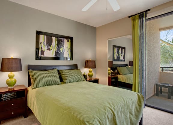 Modern bedroom here at Aspire at Pinnacle Peak Apartments with green-themed decor. Features include a queen bed, abstract artwork, matching lamps, wooden side tables, and a glass door leading to a balcony.