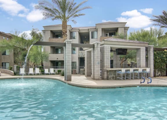 Luxurious pool area here at Aspire at Pinnacle Peak Apartments with clear blue water, lined with white lounge chairs. A modern cabana with a TV and bar seating creates a relaxed atmosphere.