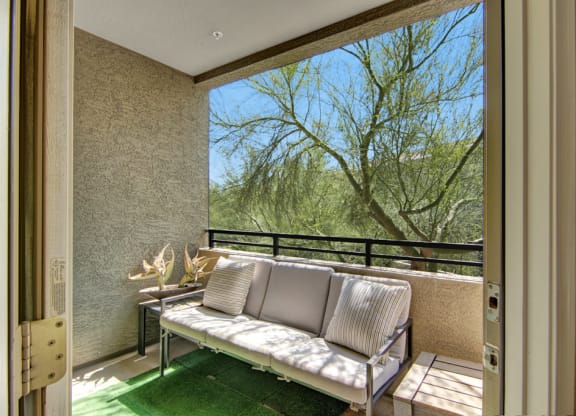 Cozy balcony with a cushioned sofa and striped pillows, overlooking a lush tree here at Aspire at Pinnacle Peak Apartments. Sunlight enhances the relaxed, natural ambiance.