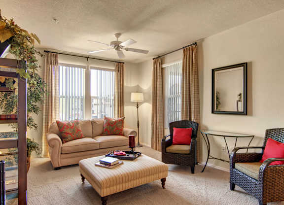A warm and inviting living room here at Bela Rosa featuring a plush sofa with accent pillows, two woven armchairs with bold cushions, and a striped ottoman coffee table. Large windows with vertical blinds and curtains fill the space with natural light, while a ceiling fan, floor lamp, and wall mirror complete the comfortable and thoughtfully styled setting.